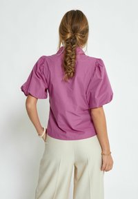 Lilla puff-sleeve pluus lühikese kaelusega, valmistatud siledast kangast, koos beežide kõrge vöökohaga pükstega. Näha on punutud juuste detail.