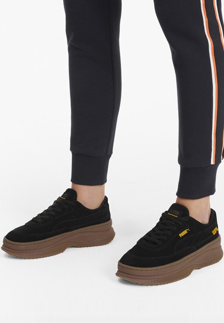 Baskets en suède noir avec une épaisse semelle en caoutchouc brun. Elles présentent des lacets noirs et des accents jaunes sur la languette et le logo latéral.