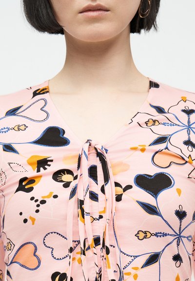 Γυναίκα που φοράει ένα ροζ ανθισμένο τοπ με σχέδια από μπλε, μαύρες και κίτρινες καρδιές και φύλλα, με έναν κόμπο μπροστά στο λαιμό.