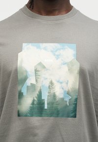 Γκρι βαμβακερό μπλουζάκι με μεγάλο γραφικό που απεικονίζει διάφορα γεωμετρικά σχήματα και σιλουέτες δάσους με φόντο έναν συννεφιασμένο ουρανό.