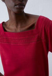 Röd topp med fyrkantig halsringning och intrikata spetsdetaljer längs axlarna. Tillverkad av mjukt material med korta ärmar.