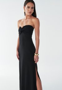 Melnie maksimālie kleita bez siksnām ar savītu mezglu detalizāciju pie krūšu daļas, sānu šķēlumu un gludu audumu. Modelī valkā zeltītu aproci un auskari.