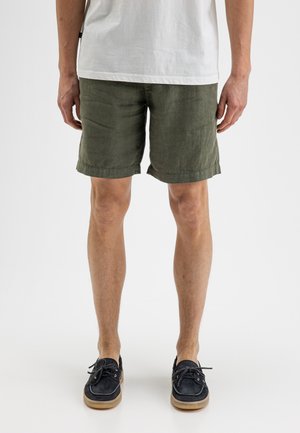 Mann steht und trägt olivgrüne Shorts, ein weißes T-Shirt und schwarze Bootsschuhe mit beigefarbenen Sohlen vor weißem Hintergrund.