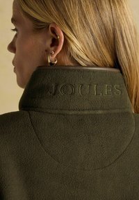 Blondynka w złotych kolczykach w kształcie kół oraz kolczykach wkrętach, widziana z tyłu, w oliwkowej zielonej kurtce polarowej z haftem "JOULES" na kołnierzu.