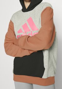 Šedá, černá a hnědá mikina s kapucí a přední kapsou ve stylu klokánka; má růžové logo Adidas a kontrastní rukávy. Vyrobená z měkké látky.