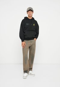 Hoodie noir avec logo sur la poitrine, casquette grise, jean délavé marron clair et baskets blanches. Le hoodie a une poche kangourou et un capuchon avec cordon de serrage.