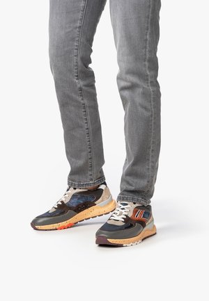 Persona che indossa jeans grigi e sneaker multicolore con suole maculate beige e arancioni, in piedi su uno sfondo bianco.