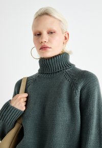 Chunky turtleneck sweater in textured dark green knit, featuring ribbed detailing and dropped shoulders, paired with a beige shoulder bag.