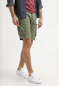 Kargo šortky v olivově zelené látce s několika postranními kapsami, kombinované s červeným tričkem a námořnickou button-up košilí. Bílé tenisky.