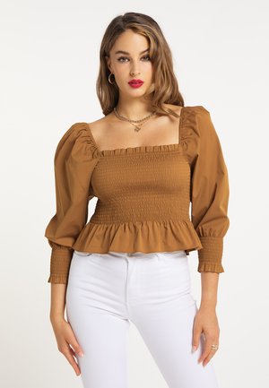 Vrouw met lichtbruin haar die een bruin gesmockt bovenstuk met pofmouwen en een witte broek draagt, staand tegen een witte achtergrond.