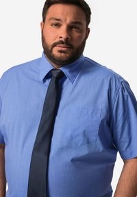 Camicia azzurra a maniche corte con colletto a punta, dotata di una tasca sul lato sinistro del petto. Abbinata a una cravatta blu scura. Tessuto liscio.