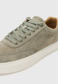Tenisi gri din suede cu sireturi verde deschis și talpă din cauciuc alb. Prezintă brandingul „Calvin Klein” embossat pe lateral. Textură netedă.