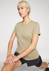 T-shirt beige ajusté avec un col rond, des manches courtes et un petit logo circulaire vert sur la poitrine gauche. Tissu doux et lisse.