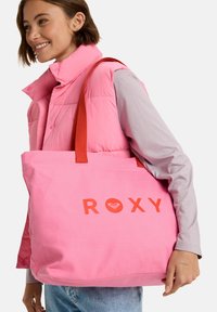 Roze canvas tote bag met rode handvatten en "ROXY" logo, met een groot hoofdcompartiment en een eenvoudig, rechthoekig ontwerp.