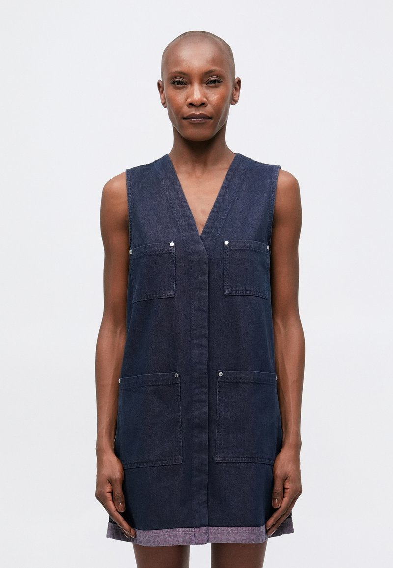 Denim brezrokavnik obleka v temno modri barvi z V-izrezom, dvema sprednjima žepoma in robom, ki ga poudarja svetlejša vijolična črta.