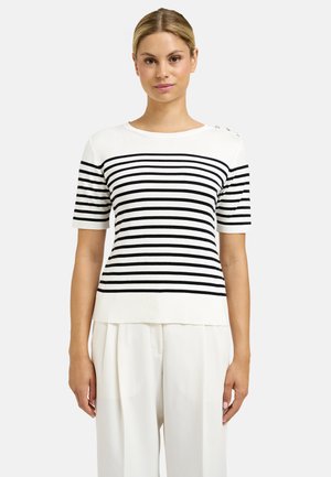 Woman with blonde hair wears short-sleeve white sweater with black horizontal stripes and white high-waist pants, standing against plain background.