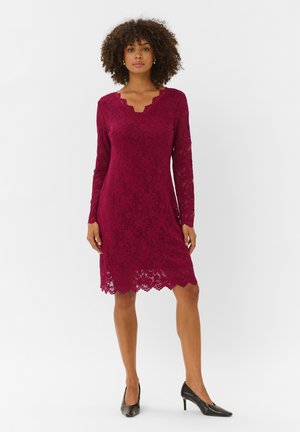 Femme debout portant une robe en dentelle bordeaux à manches longues et longueur genou, ainsi que des escarpins noirs à bouts pointus sur un fond uni.