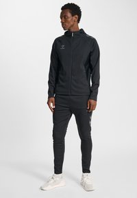 Zwart atletisch trainingspak met jas met ritssluiting en taps toelopende broek; voorzien van reflecterende details en een logo. Draagt lichte sneakers.