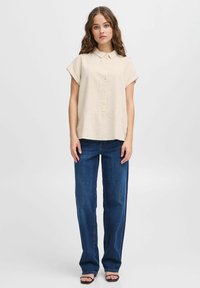 Camicia beige a maniche corte con colletto e chiusura frontale a bottoni, abbinata a jeans a vita alta blu scuro a gamba larga. Tessuto morbido, vestibilità rilassata.
