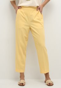 Un pantalon jaune ajusté au toucher lisse, avec une taille haute, des poches latérales et des jambes fuselées. Associé à des sandales plates beige.