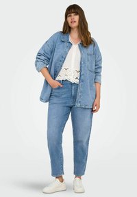Veste en jeans bleu portée sur un haut blanc à dentelle, associée à un jean bleu clair et des baskets blanches. Tenue décontractée avec une coupe relax.