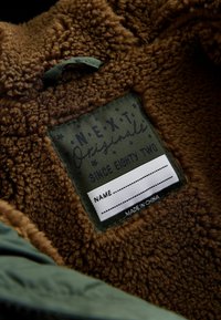 Brun fleeceforing med en grøn stofmærkat, der har teksten "NEXT Originals Since Eighty Two" og et tomt navnfelt. Fremstillet i Kina.