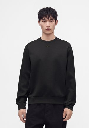 Giovane uomo con capelli corti neri che indossa una felpa nera semplice e pantaloni neri, in piedi davanti a uno sfondo bianco.