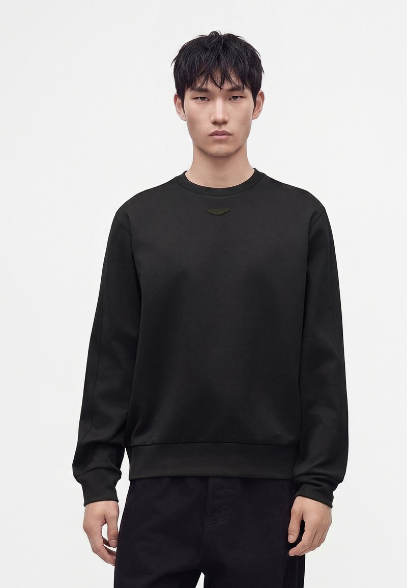 Jeune homme aux cheveux courts noirs portant un sweat-shirt noir uni et un pantalon noir, debout devant un fond blanc.