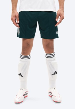 Apatinė žmogaus kūno dalis, dėvinti juodus Adidas sportinius šortus, baltas kelių aukščio Adidas kojines su blauzdų apsaugomis ir baltus Adidas futbolo batelius.