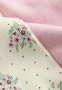 Krémová látka s ribbenou texturou, která má růžové květinové vzory a hnědé tečky, s vrstvou nad hladkým pastelově růžovým materiálem.