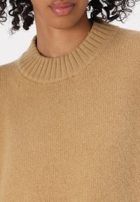 Maglione beige a maglia con colletto a girocollo a costine e una morbida texture, in primo piano sui dettagli del tessuto e delle cuciture.