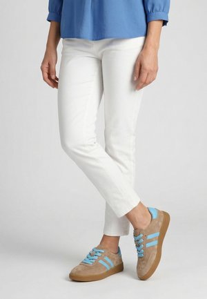 Persoon draagt witte slim-fit broek, een blauw lang mouw shirt en beige sneakers met lichtblauwe veters en strepen, staand met gekruiste benen.