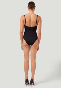 Svart baddräkt i ett stycke med justerbara axelband, medelcut rygg, slät textur och minimalistisk design, tillsammans med svarta högklackade skor.