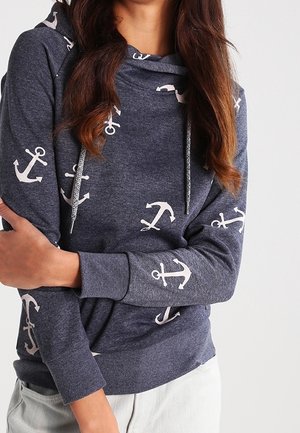 Vrouw draagt een marineblauwe hoodie met wit ankerpatroon en trekkoorden, armen gekruist, staande tegen een effen lichte achtergrond.
