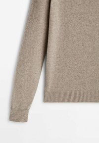 Manche de pull en maille beige et ourlet inférieur avec poignets et bord inférieur côtelés sur fond blanc.
