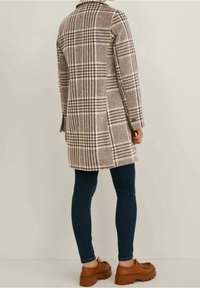 Abrigo marrón a cuadros con cuello, de mangas largas y aberturas laterales, hecho de un tejido texturizado. Combinado con jeans azul oscuro y zapatos marrones.