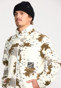 Veste en polaire crème avec des taches marron et gris, col haut, fermeture éclair à l'avant et une poche poitrine. Bonnet en tricot noir porté.