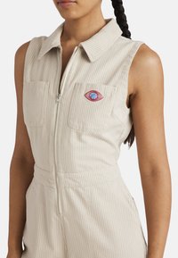 Mono de rayas beige con cuello, cierre frontal y dos bolsillos en el pecho. Presenta un diseño bordado de un ojo en rojo y azul en el bolsillo izquierdo.