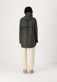 Olijfgroene regenjas met een capuchon en een flap aan de achterkant, gecombineerd met off-white broeken en beige schoenen. Glad stof, relaxte pasvorm.