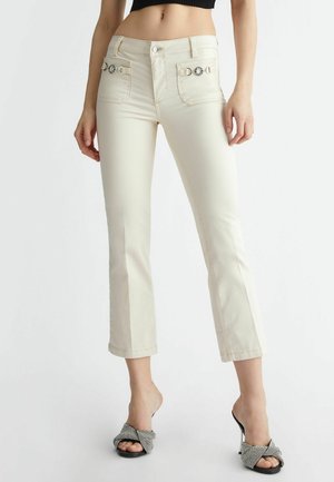 Kobieta ubrana w beżowe spodnie o wysokim stanie, krótkie, z przednimi kieszeniami i srebrnymi guzikami, zestawione z czarno-białymi sandałami na obcasie.