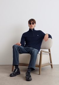 Pulover cu fermoar bleumarin, blugi, pantofi negri și ochelari de soare. Modelul stătea pe un fotoliu ușor cu picioare din lemn, în fața unui perete simplu.