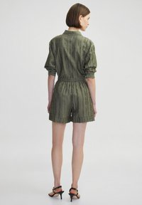 Salopette corta verde oliva, texturizzata, con maniche corte, vita elastica e colletto. Indossata con tacchi neri con cinturini.