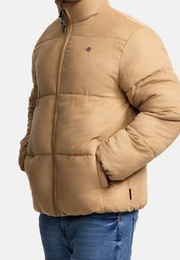 Beżowa ocieplana kurtka z wysokim kołnierzem, zamek błyskawiczny z przodu i bocznymi kieszeniami. Miękka tekstura, pikowany fason oraz subtelne logo na piersi.