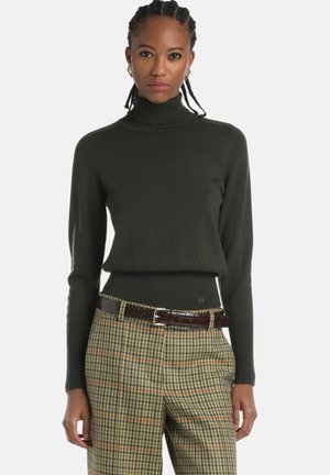 Donkergroene coltrui met ribbeltextuur, voorzien van een getailleerde taille. Gepaard met een klassieke broek in aardetinten, geaccentueerd met een bruine riem.