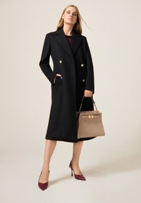 Cappotto doppiopetto nero con bottoni dorati, abbinato a una borsa beige chiaro. Modella che indossa un top bordeaux e décolleté borgogna a punta.