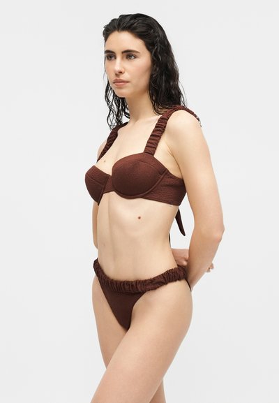 Femme aux cheveux mouillés portant un bikini marron foncé texturé avec des bretelles froncées, debout les mains derrière le dos sur un fond uni.