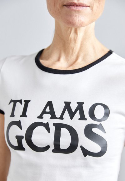 T-shirt branca com texto preto a dizer "TI AMO GCDS," apresentando um decote redondo e um acabamento preto em contraste nas mangas e na gola.