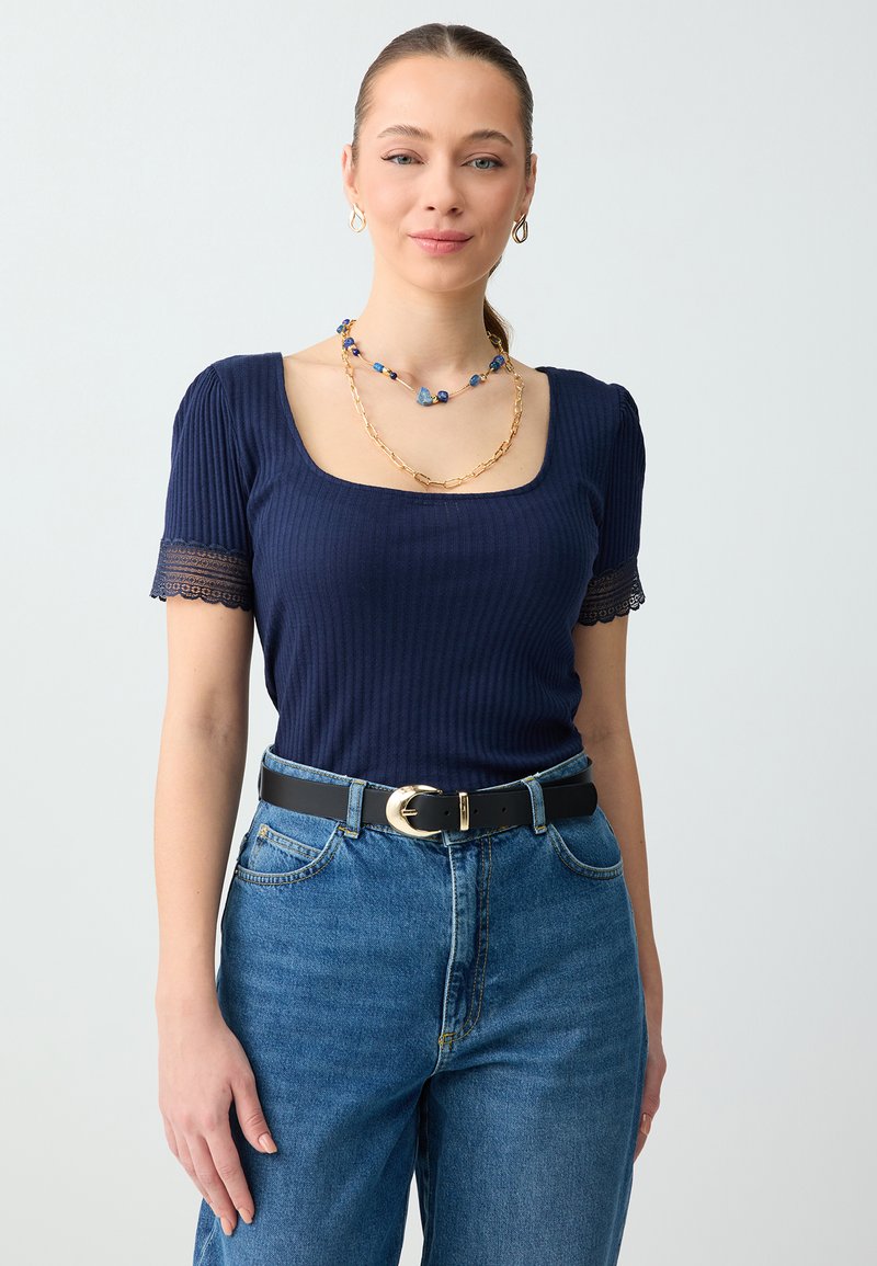 Femme portant un haut côtelé bleu marine avec des manches en dentelle, un jean bleu, une ceinture noire avec une boucle dorée, et des colliers superposés avec des perles bleues et une chaîne dorée.
