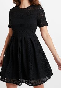 Schwarzes, kurzärmliges Kleid aus strukturiertem Spitze-Material. Es hat ein tailliertes Oberteil und einen ausgestellten Rock, mit einem gemusterte Saumlinie und rundem Ausschnitt.