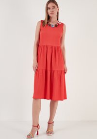 Rotes, ärmelloses Kleid aus strukturiertem, geripptem Gewebe. Hat ein gestuftes Design und eine bunte Halskette am Ausschnitt. Knielang.
